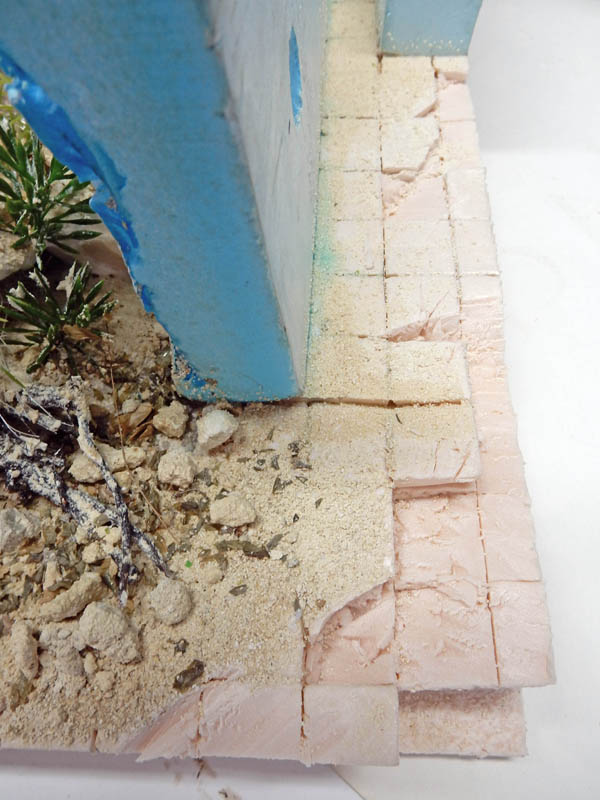 Sol pavé de maquette texturé avec sable, débris et végétation contre un mur bleu