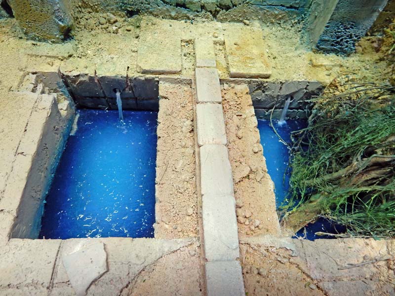 Bassins d'eau bleue d'une maquette de lavoir avec margelles en pierre et végétation