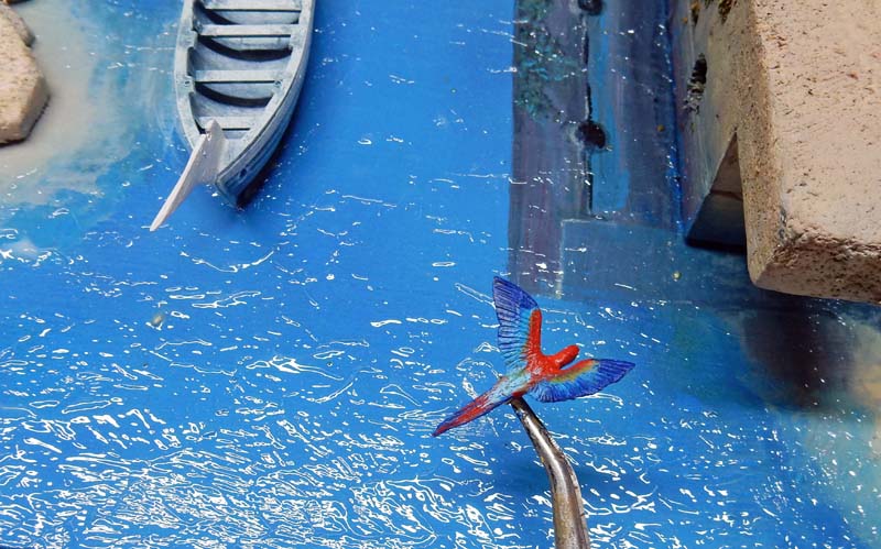 Martin-pêcheur miniature peint tenu à la pince au-dessus d'une maquette d'eau bleue avec barque