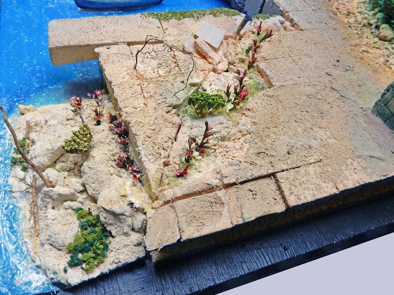 Diorama de maquettisme représentant un terrain rocheux fleuri bordant une eau bleue