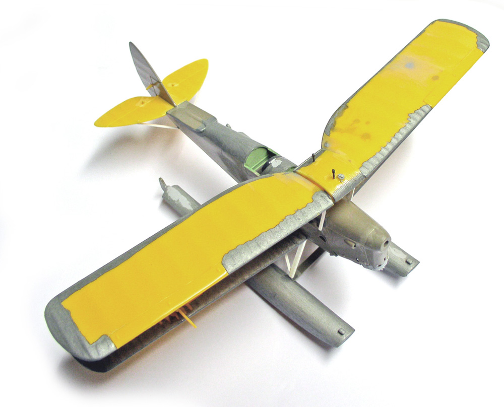 Maquette d'hydravion à flotteurs en cours de peinture, ailes jaunes sur fuselage gris, fond blanc