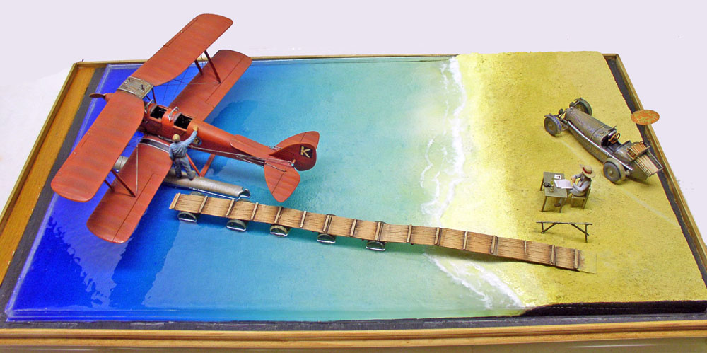 Diorama d'hydravion biplan rouge amarré près d'un ponton, plage et véhicule militaire