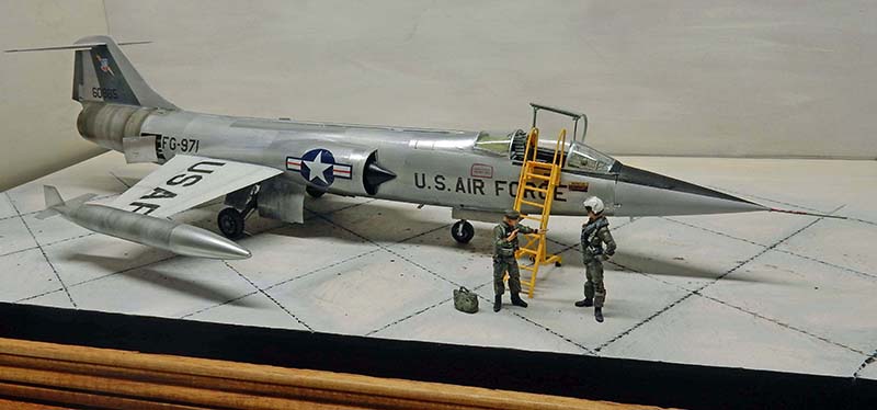 Diorama d'un F-104 Starfighter argenté sur tarmac avec échelle jaune et deux pilotes