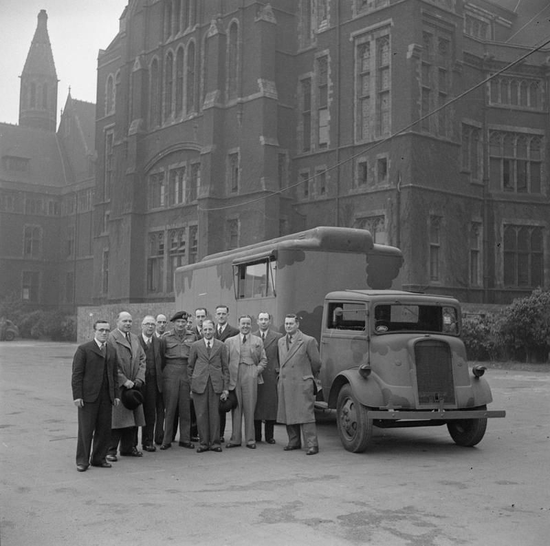 Photo ancienne en noir et blanc d'un camion Fordson 7V entouré d'hommes en costume devant un grand bâtiment