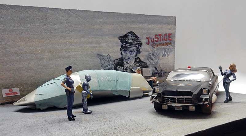 Diorama futuriste avec policière, robot, voiture de police noire et figurine féminine devant un mur graffé