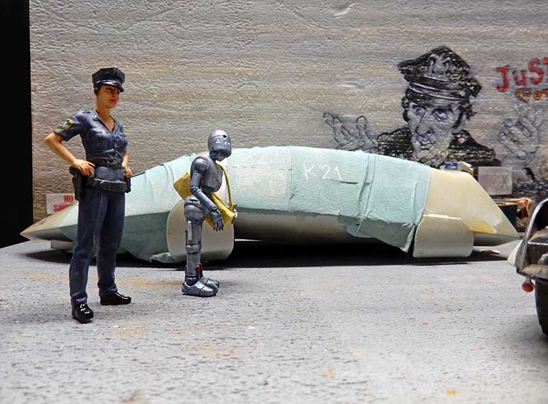 Diorama avec figurine de policière et robot androïde devant une épave près d'un mur graffité