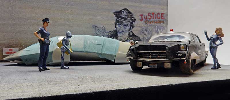 Diorama futuriste avec policière, robot, voiture noire et personnage féminin devant un mur graffé « Justice »