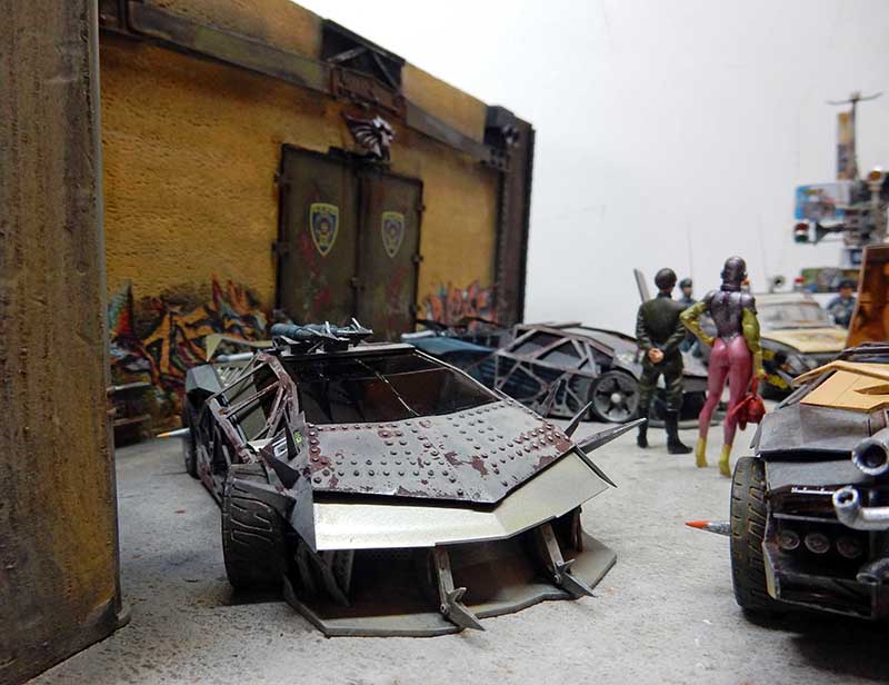 Maquette de voiture blindée post-apocalyptique avec figurines devant un mur tagué et portail métallique