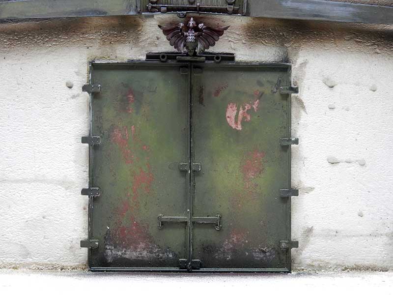 Porte métallique blindée vert patiné avec traces rouges, encastrée dans un mur blanc