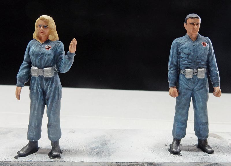 Deux figurines peintes en combinaison bleue, une femme blonde et un homme, sur fond noir