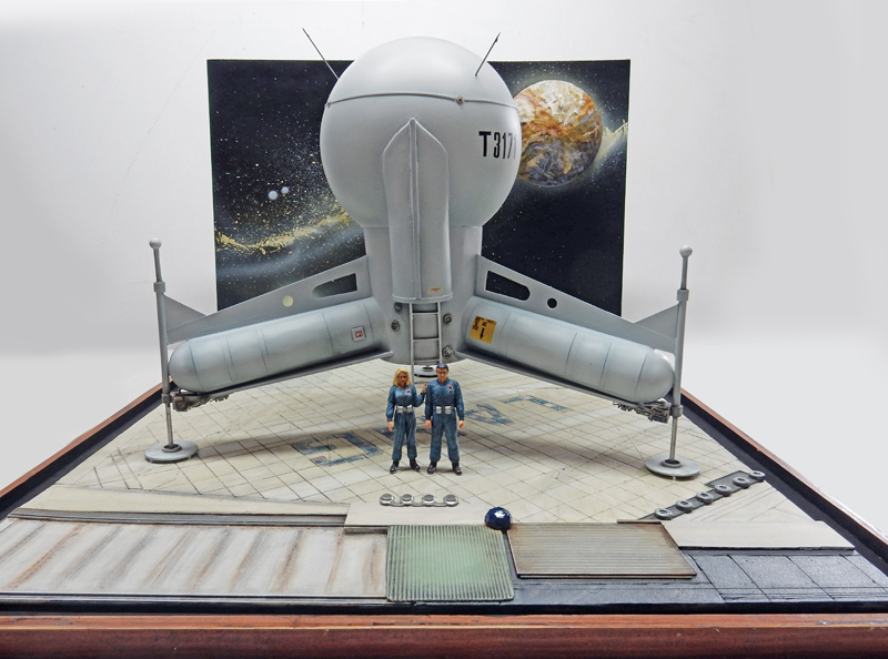 Maquette du G.N.U. argenté posé sur sa base avec deux figurines devant un décor spatial