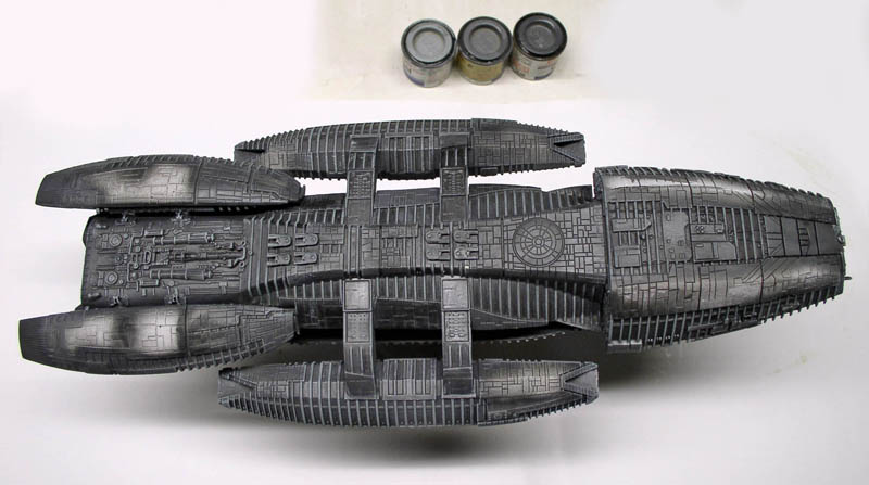 Maquette grise du vaisseau Battlestar Galactica vue de dessus avec trois pots de peinture