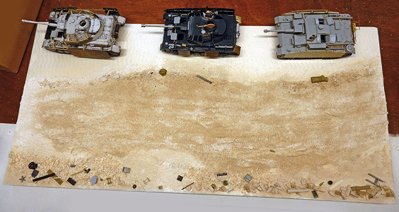 Trois maquettes de chars allemands aux finitions différentes posées sur un diorama de terrain sablonneux en préparation