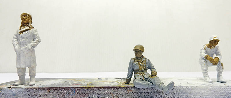 Trois figurines de soldats au 1/35e en cours de peinture, dont un assis avec casque
