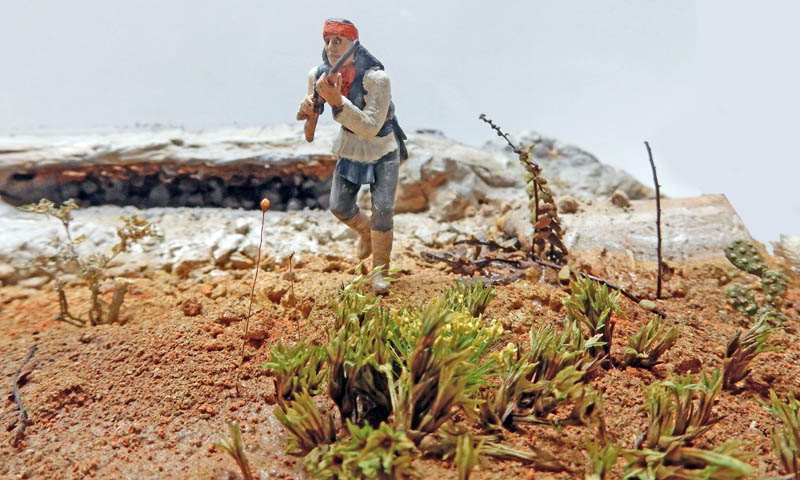 Figurine d'Indien apache au bandeau rouge avançant fusil en main sur un diorama désertique