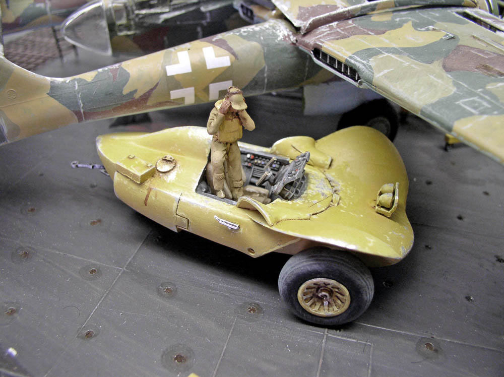 Maquette d'engin jaune au pilote debout assemblé par kitbash devant un avion militaire camouflé