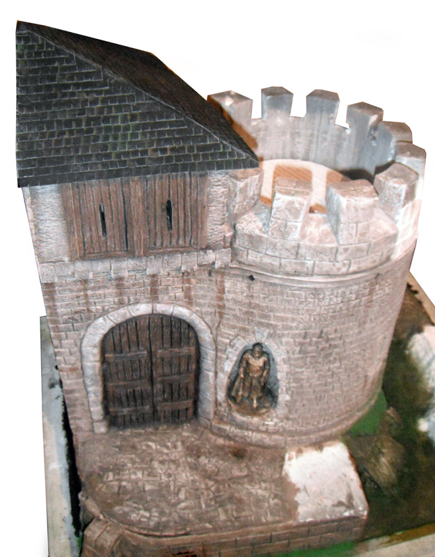 Maquette de donjon médiéval avec tour crénelée, porte en bois et figurine de garde