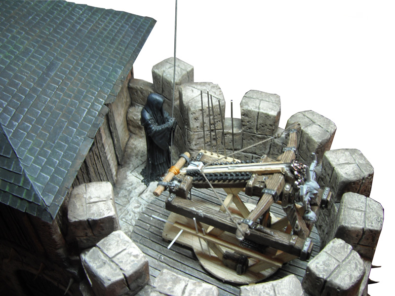 Maquette du sommet d'un donjon crénelé avec catapulte et figurine encapuchonnée sur plancher de bois
