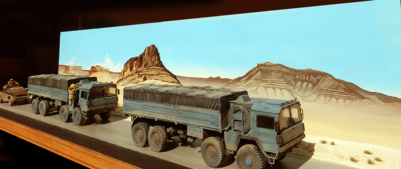 Diorama de deux camions militaires bâchés traversant un désert montagneux devant un piton rocheux