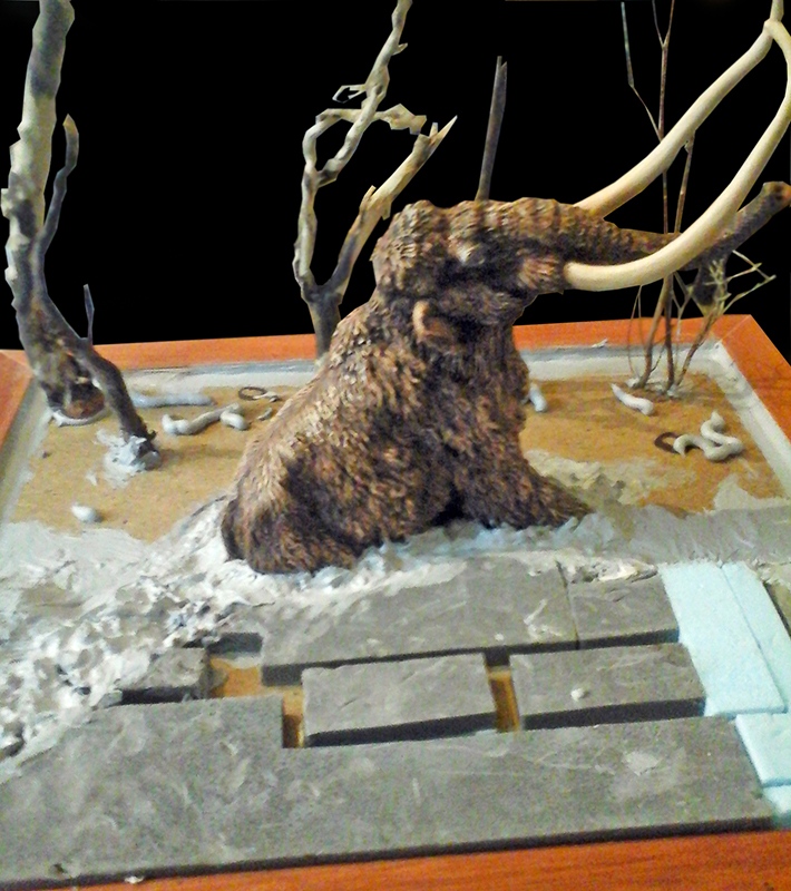 Maquette de mammouth laineux enlisé dans un diorama avec arbres morts et neige