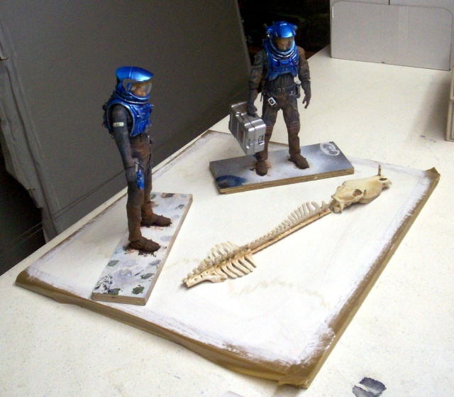 Deux figurines d'astronautes en combinaison bleue près d'un squelette d'animal sur socles en cours de fabrication
