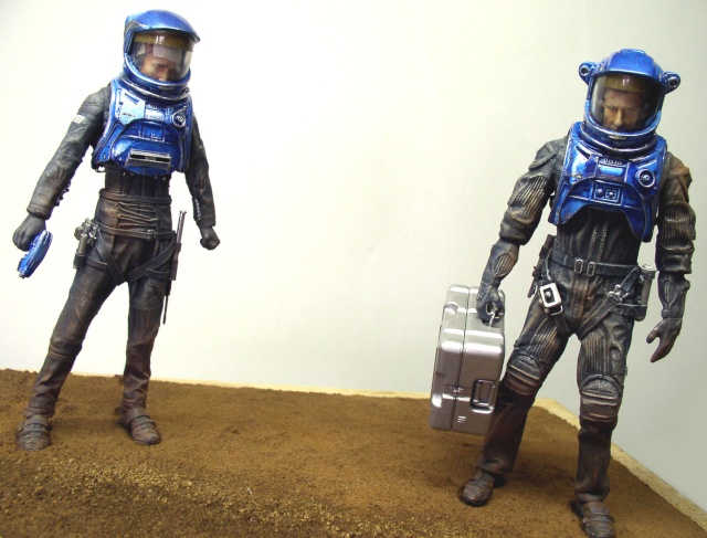 Deux figurines d'astronautes en combinaison bleue marchant sur un sol martien, l'une portant une mallette argentée