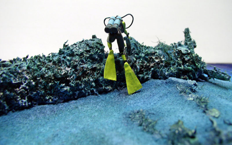 Figurine de plongeur aux palmes jaunes explorant l'épave immergée et envasée du Mitsubishi A6M5