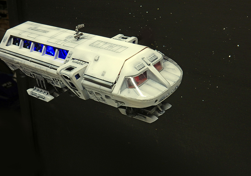 Maquette du Moonbus blanc aux hublots bleus éclairés posée sur fond étoilé noir