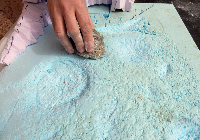 Main sculptant des cratères dans une plaque de mousse bleue avec une pierre pour le diorama lunaire