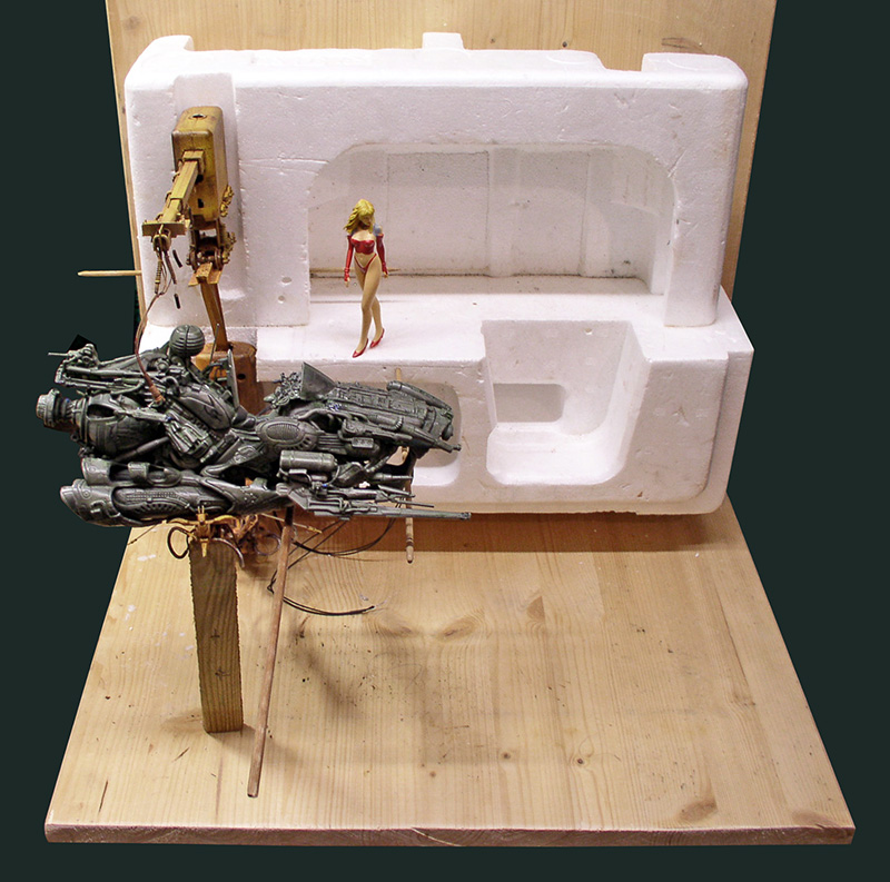 Maquette de motovolante mécanique métallisée et figurine féminine devant un support en polystyrène
