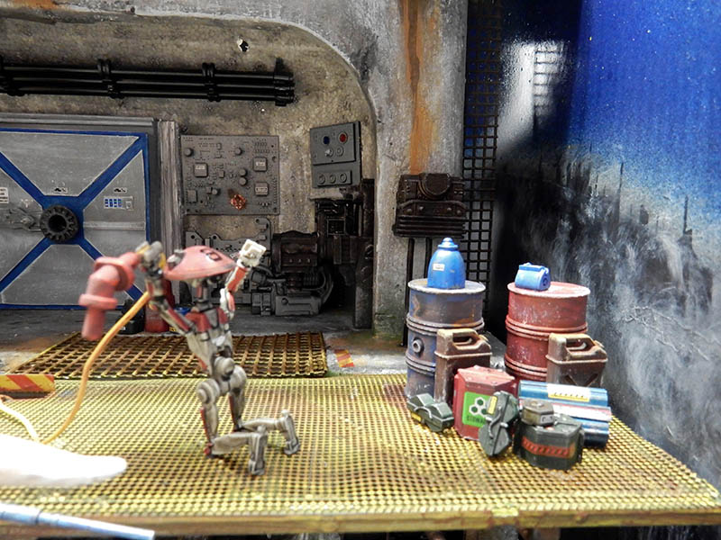 Diorama de hangar industriel avec robot rouge agenouillé, bidons rouillés et caisses sur plancher métallique