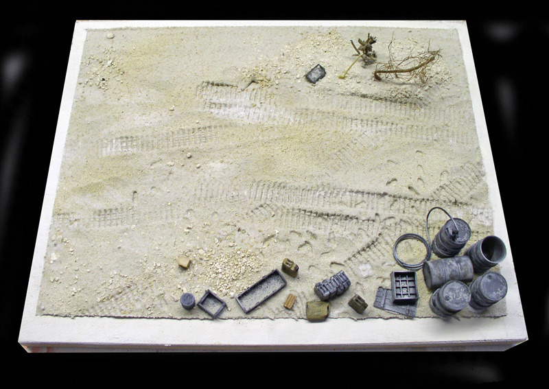 Socle de diorama sablonneux avec traces de chenilles, bidons, caisses et accessoires miniatures avant peinture