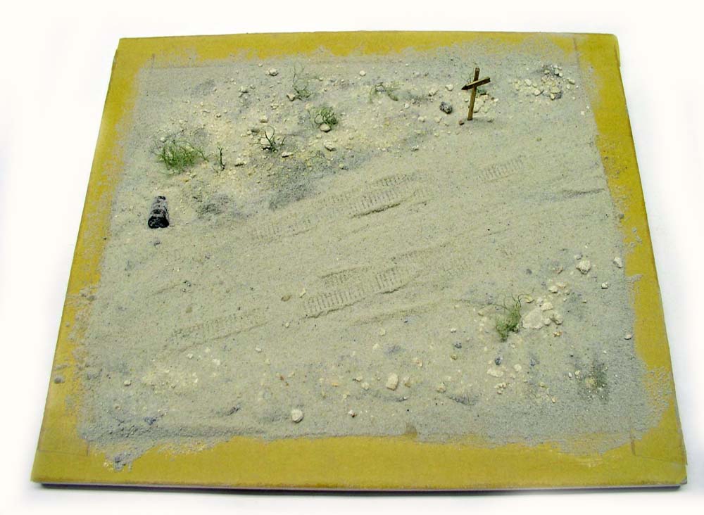Socle de diorama désertique avec traces de chenilles, touffes d'herbe et petite croix en bois