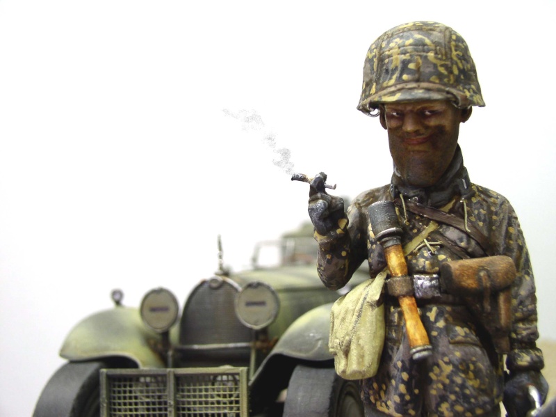 Figurine peinte de soldat fumant en tenue camouflée devant une maquette de véhicule militaire