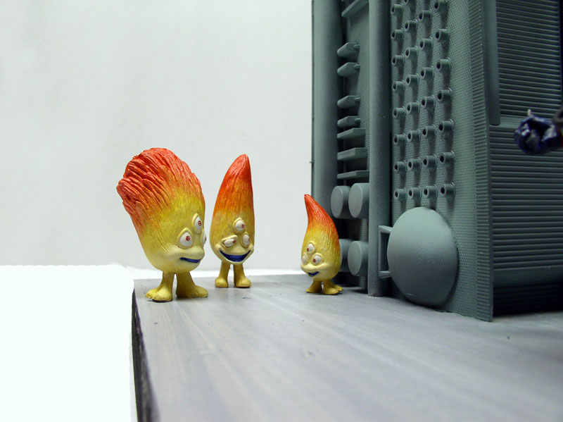 Trois figurines de personnages flamme jaune orangé devant une structure mécanique grise en maquette