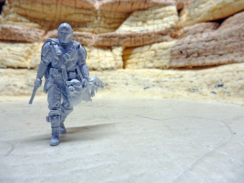 Figurine grise du Mandalorien portant Grogu marchant dans un décor désertique rocheux