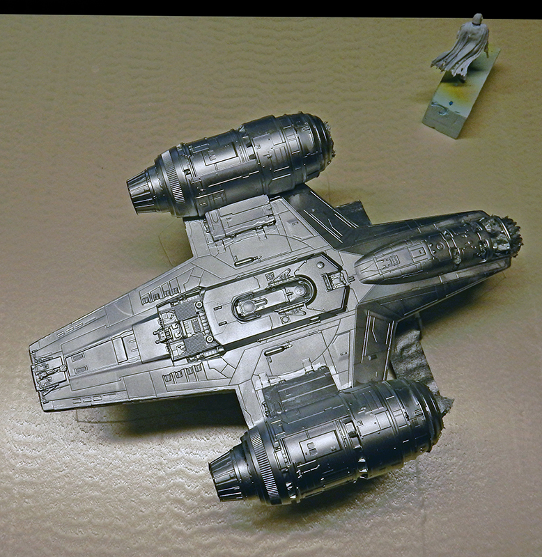 Maquette du Razor Crest peinte en gris vue de dessus avec figurine du Mandalorien en arrière-plan
