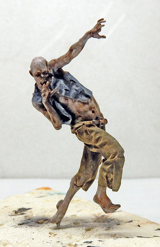 Figurine peinte de zombie décharné en pantalon déchiré, bras levé, sur socle clair