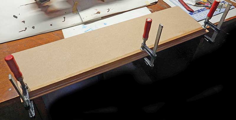 Planche de socle en MDF maintenue par des serre-joints rouges pendant le collage du Rodger Young