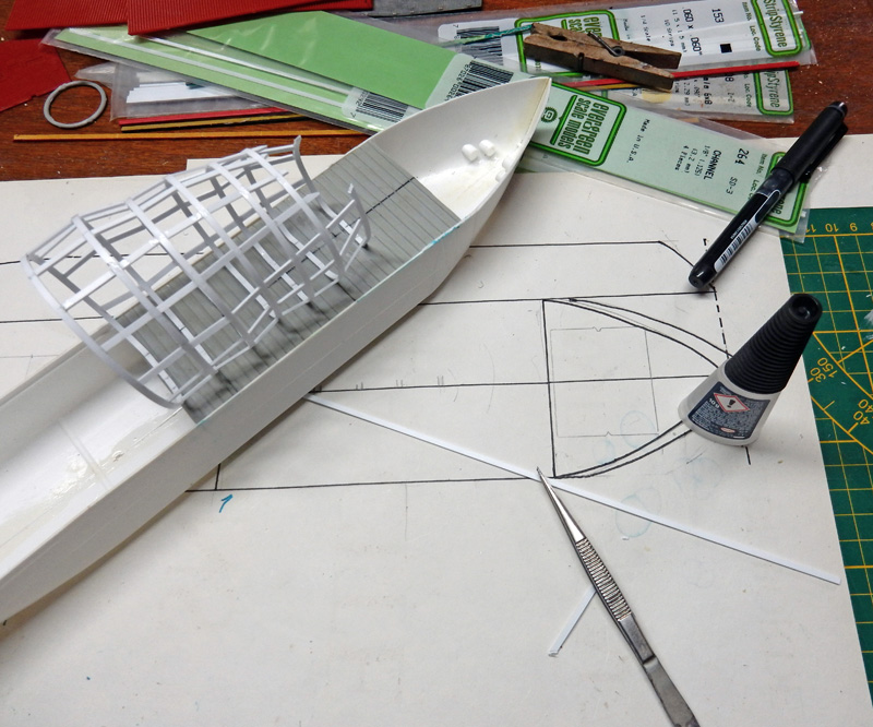 Coque de bateau en plastique blanc en construction sur plan, entourée de baguettes Evergreen et outils de modélisme