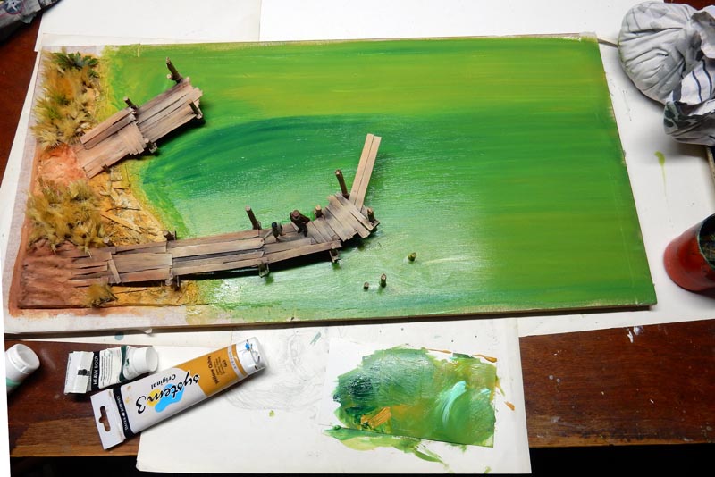 Maquette de ponton en bois sur eau verte peinte, avec tubes de peinture et palette d'artiste