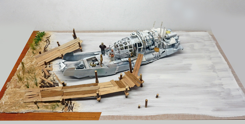 Diorama de maquette représentant une épave de bateau échouée près d'un ponton en bois avec personnages