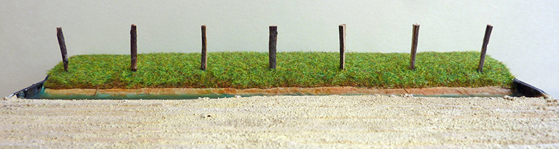 Maquette d'un terrain herbeux bordé de piquets de bois alignés sur socle sablonneux