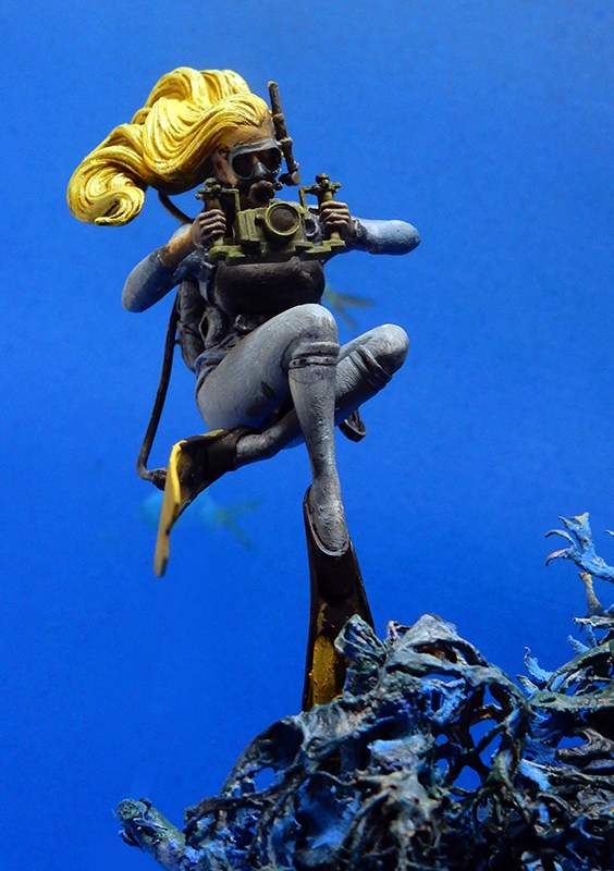 Figurine de plongeuse blonde avec appareil photo sous-marin nageant au-dessus de coraux