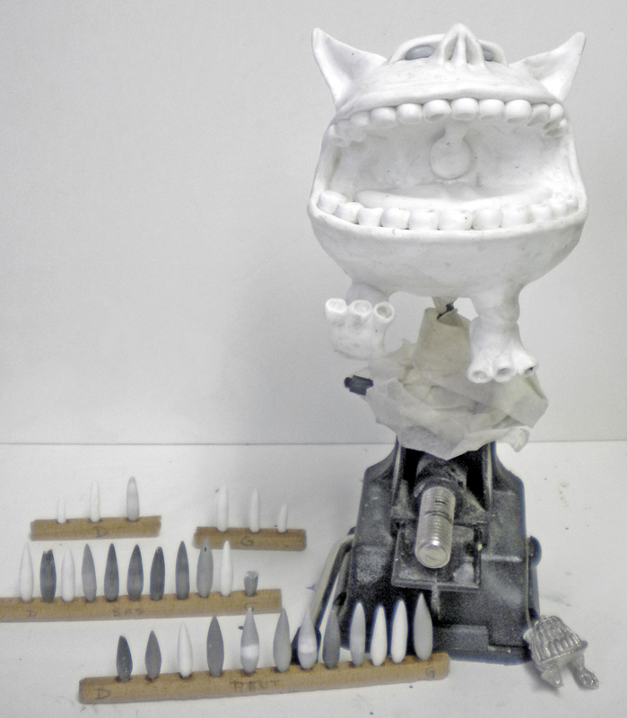 Sculpture blanche d'un Squig à grande gueule dentée en cours, entourée de dents séchant sur supports