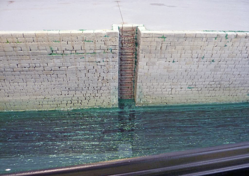Quai en pierre avec échelle rouillée surplombant une eau verte, décor de diorama