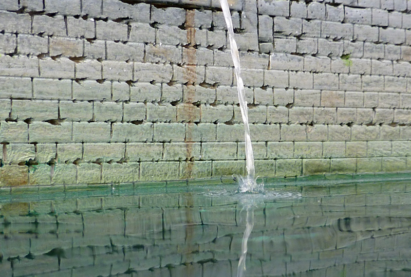 Filet d'eau tombant dans un bassin, reflets sur l'eau devant un mur de briques
