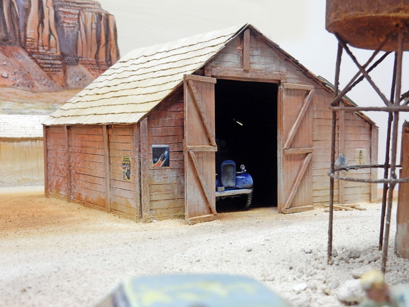 Grange en bois d'un diorama désertique abritant un véhicule bleu, château d'eau rouillé à droite