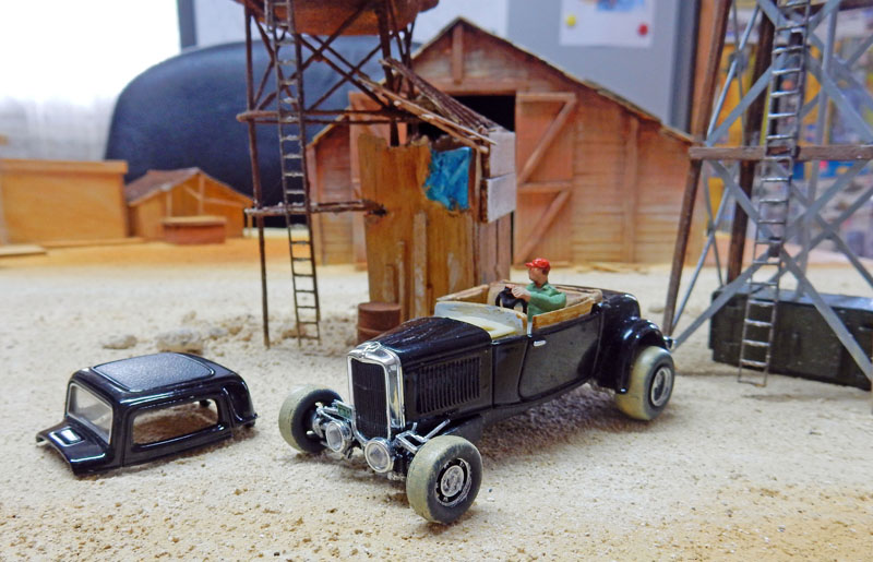 Hot rod noir miniature avec figurine sur un diorama désertique, hangar en bois et pylônes métalliques