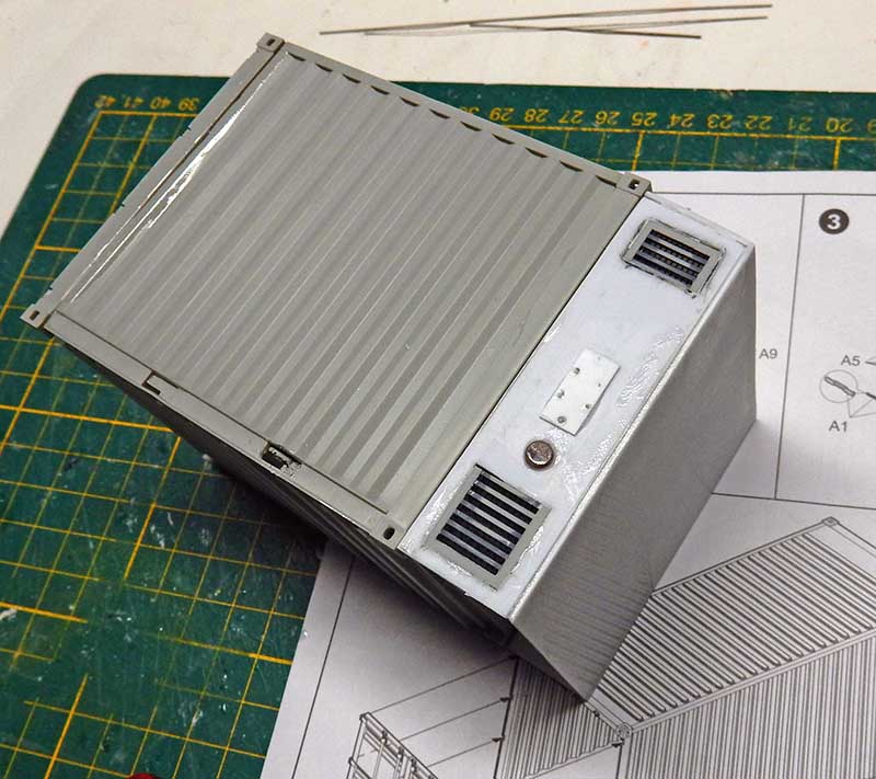 Maquette de conteneur gris avec grilles d'aération posée sur un tapis de découpe à côté de la notice